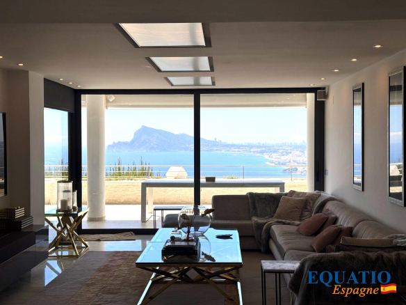 Modern living room with large glass doors opening to a sea view, a light-gray sectional sofa, and a glass coffee table overlooking the coastline.