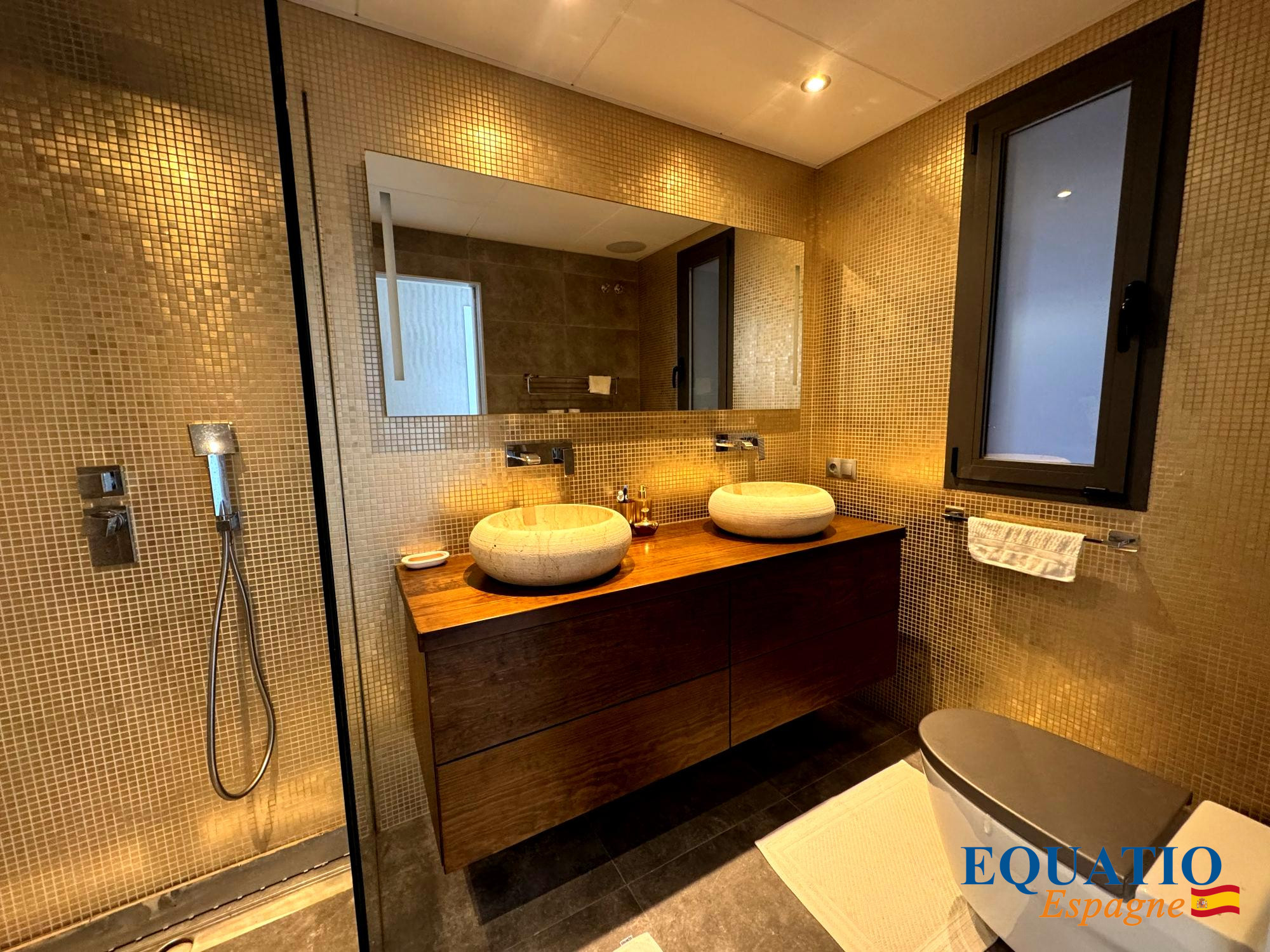 Bathroom with a wooden double vanity, two round stone vessel sinks, mosaic tile walls, a glass shower, and a toilet.