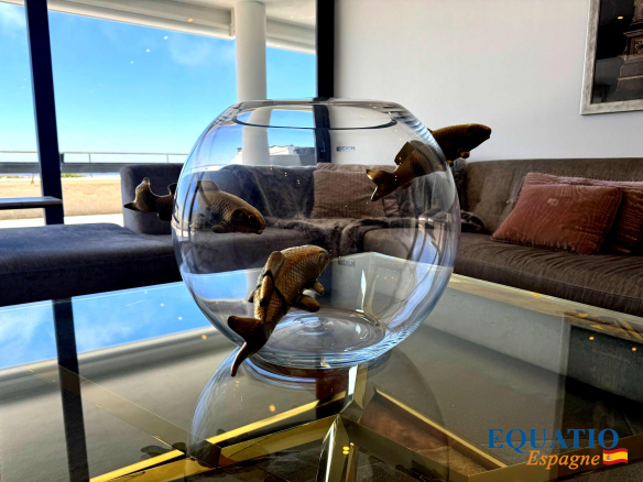 Glass bowl with three goldfish figurines, placed on a glass coffee table in a bright modern living room with a grey sofa and pink cushions.