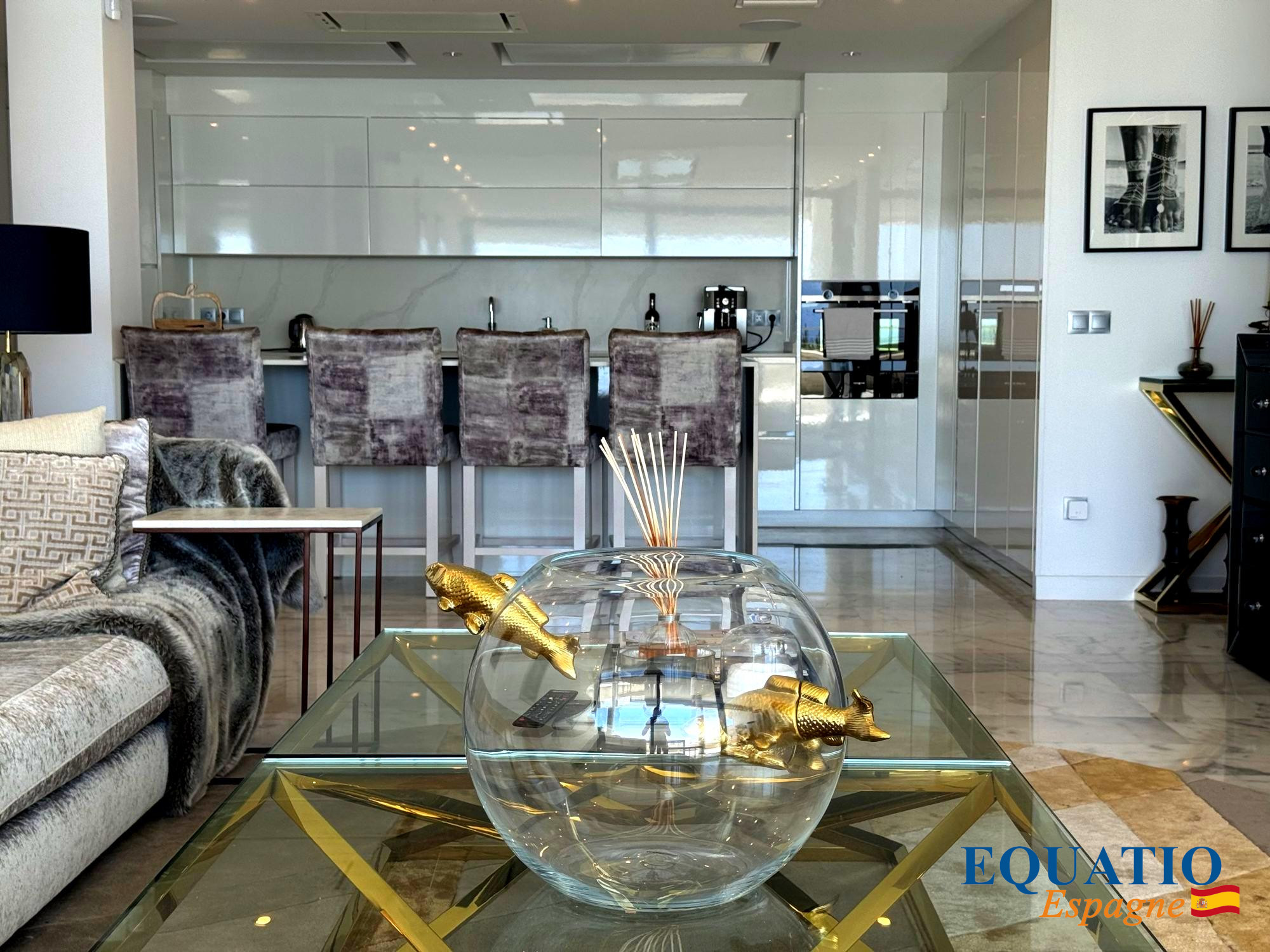Open-plan living area with a white glossy kitchen/bar in the background, four purple velvet bar stools, a plush grey sofa, and a glass coffee table featuring a large glass bowl with gold koi fish figurines in the foreground. Watermark reads 'EQUATIO Espagn e' in the bottom right.