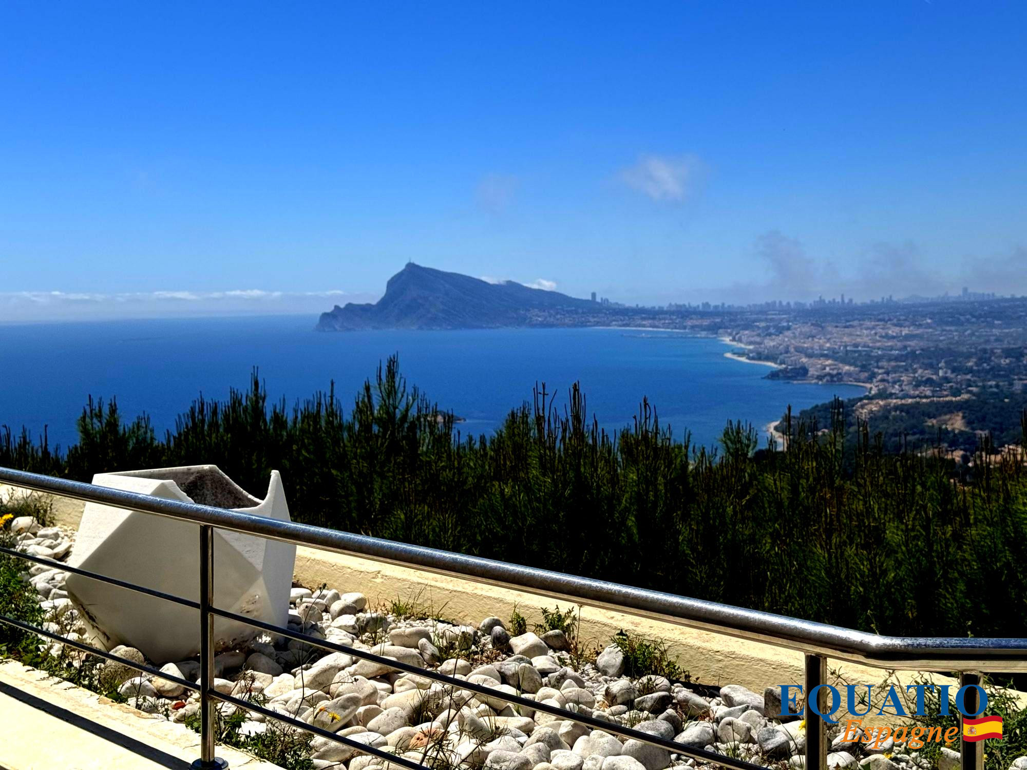 Panoramic coastal view from a lookout: rocky foreground, metal railing, and a blue sea meeting a distant city along the shore under a clear sky.