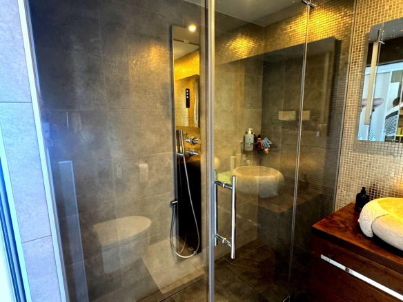 Glass-walled walk-in shower with gray tile, chrome door handle, and a handheld shower fixture; reflection of a sink and mirror in the back wall.