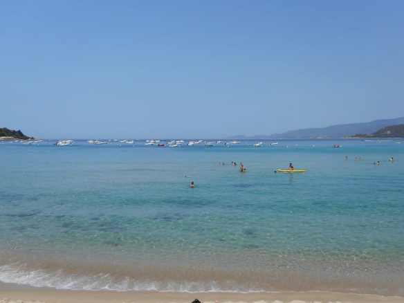 Plage de Calcatoggio avec des baigneurs et des bateaux au large, ciel dégagé.