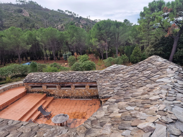 Vue de la toiture en pierre d'une bastide avec terrasse et forêt en arrière-plan.