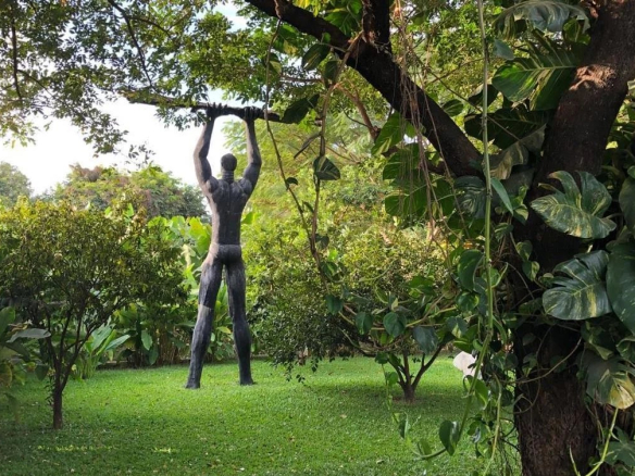 Statue en métal représentant un homme levant un bâton, entourée de verdure dans un jardin.