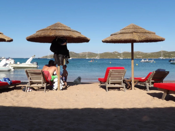 Beach with parasols and sun loungers, view of the sea and boats in the distance.