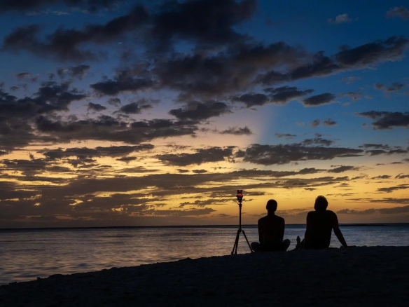 Deux personnes assises sur la plage regardant un coucher de soleil avec un trépied à proximité.