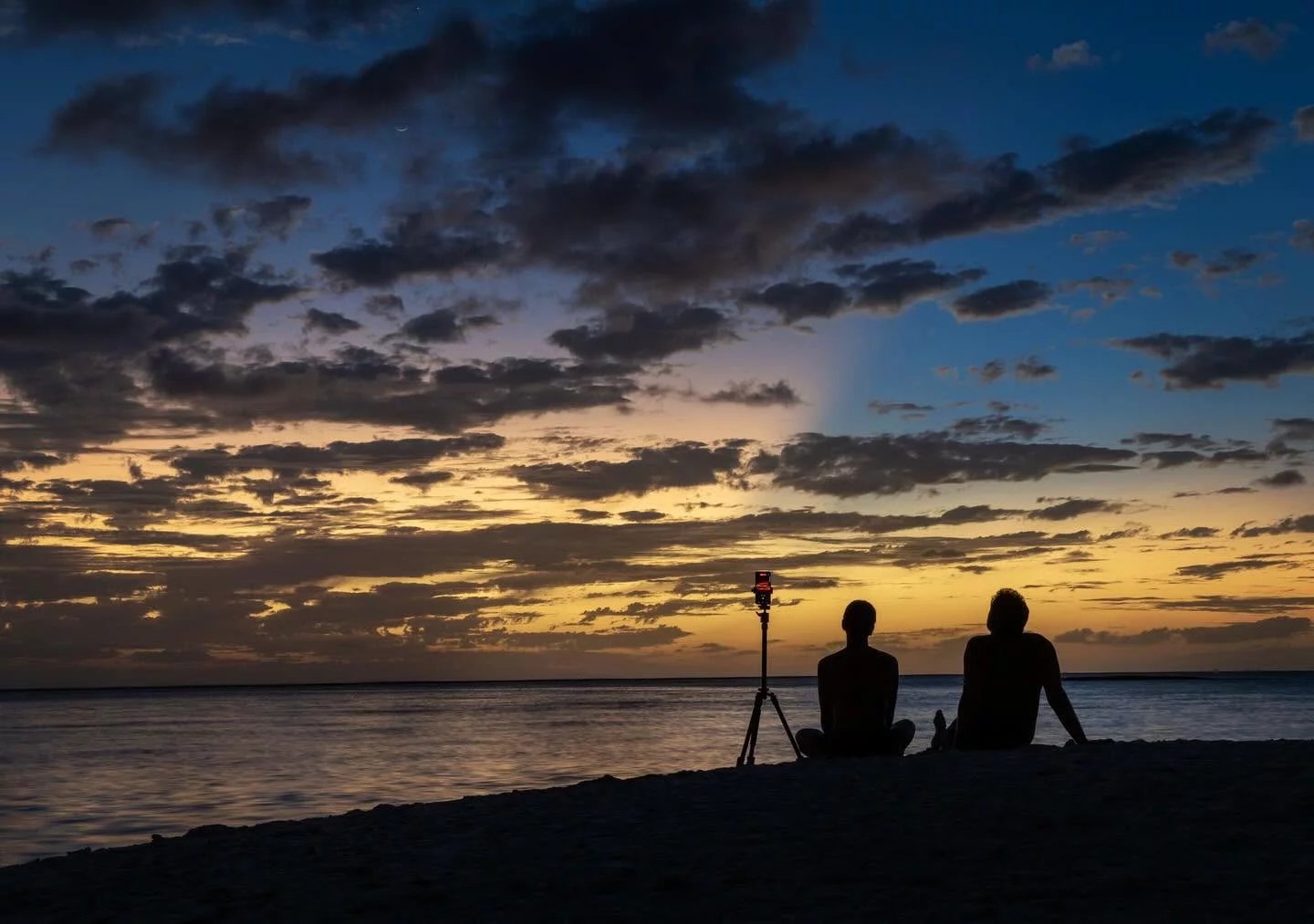 Coucher de soleil à Tikehau Deux personnes assises sur la plage regardant un coucher de soleil avec un trépied à proximité.