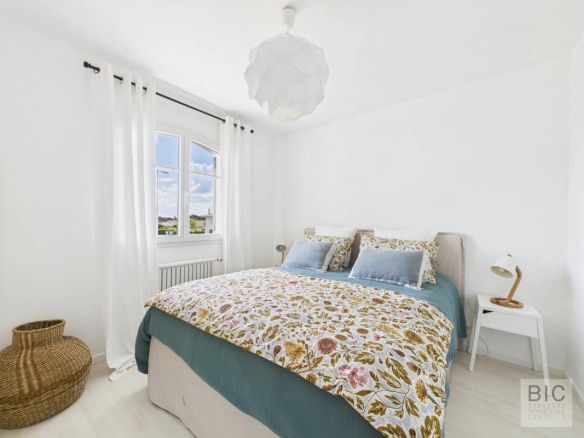 Chambre lumineuse avec lit double et décoration florale, murs blancs et fenêtres avec rideaux.