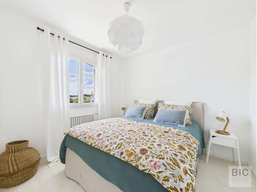 Chambre lumineuse avec lit double et décoration florale, murs blancs et fenêtres avec rideaux.