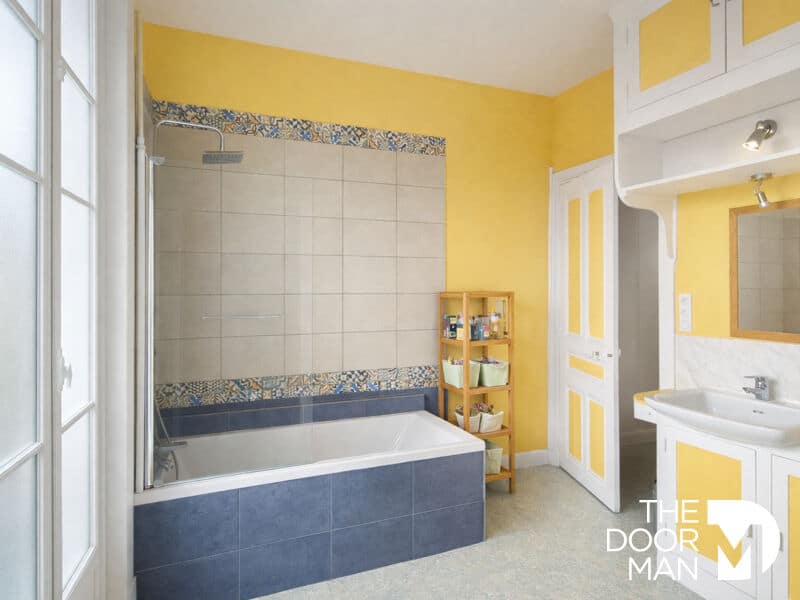 Salle de bains de maison à Rambouillet Salle de bains avec baignoire, carrelage bleu et murs jaunes, étagère en bois avec produits de toilette.
