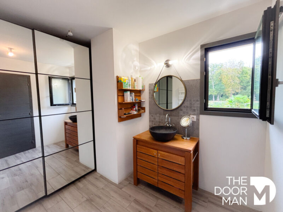 Modern bathroom with wooden furniture and round mirror, natural light coming in through a window.