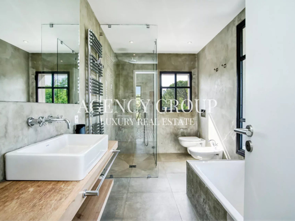 Contemporary bathroom with concrete walls, glass shower enclosure, white rectangular sink on a wooden vanity, and a freestanding bathtub under large windows.