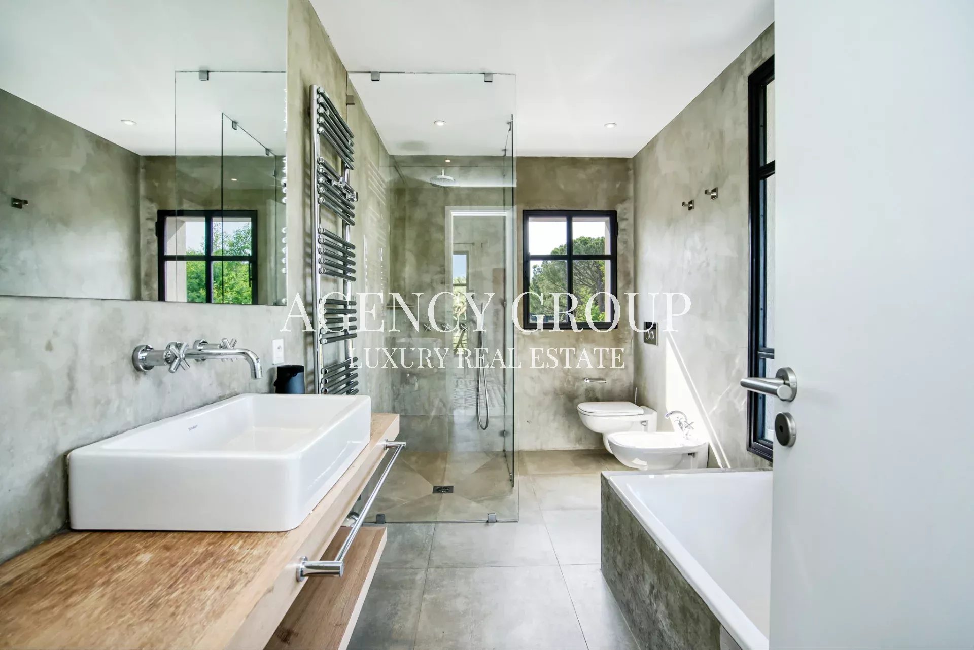 Contemporary bathroom with concrete walls, glass shower enclosure, white rectangular sink on a wooden vanity, and a freestanding bathtub under large windows.