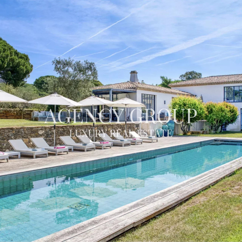 Luxurious villa with a rectangular pool, wooden deck, and white lounge chairs under umbrellas.