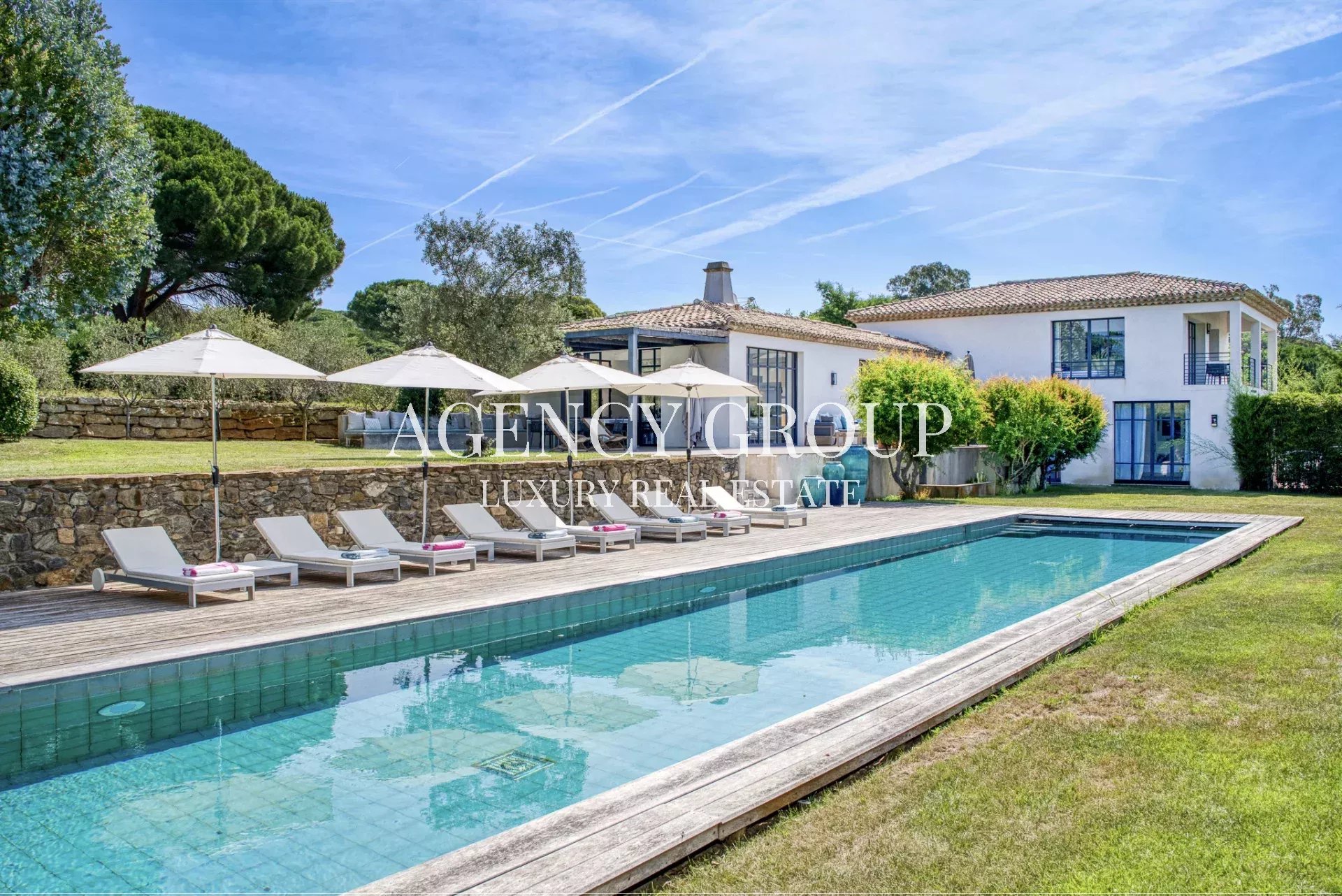 Luxurious villa with a rectangular pool, wooden deck, and white lounge chairs under umbrellas.