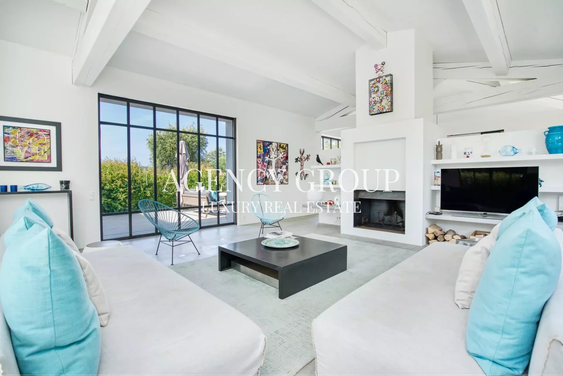 Bright, open-concept living room with white walls, large glass doors to a garden, and a black square coffee table between white sofas with turquoise cushions.