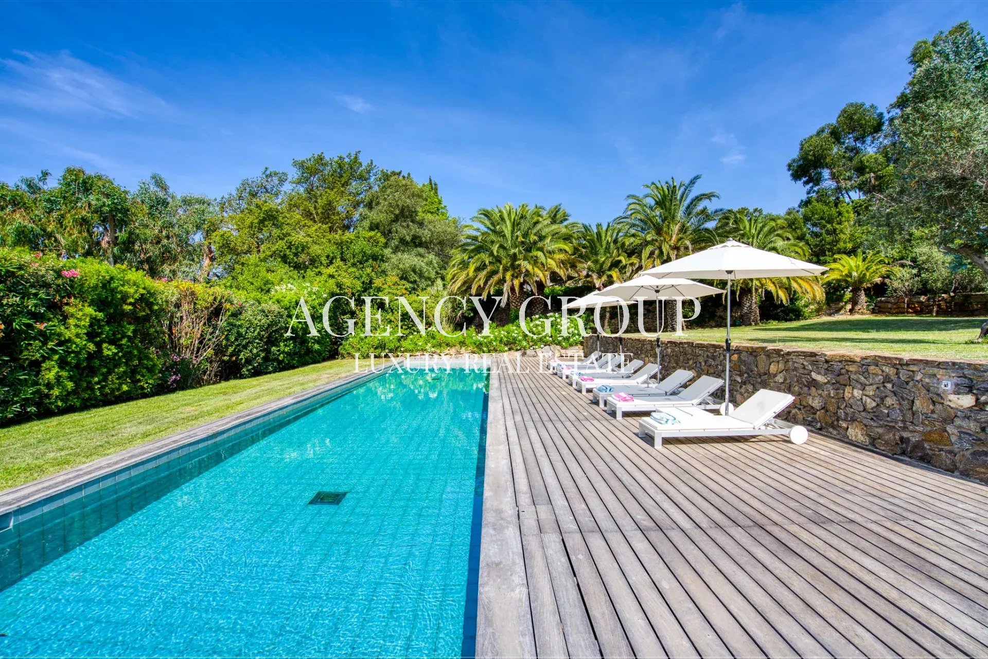 Sunny backyard pool area with a long turquoise pool, wooden deck, white lounge chairs, and large umbrellas surrounded by tropical greenery and palm trees.