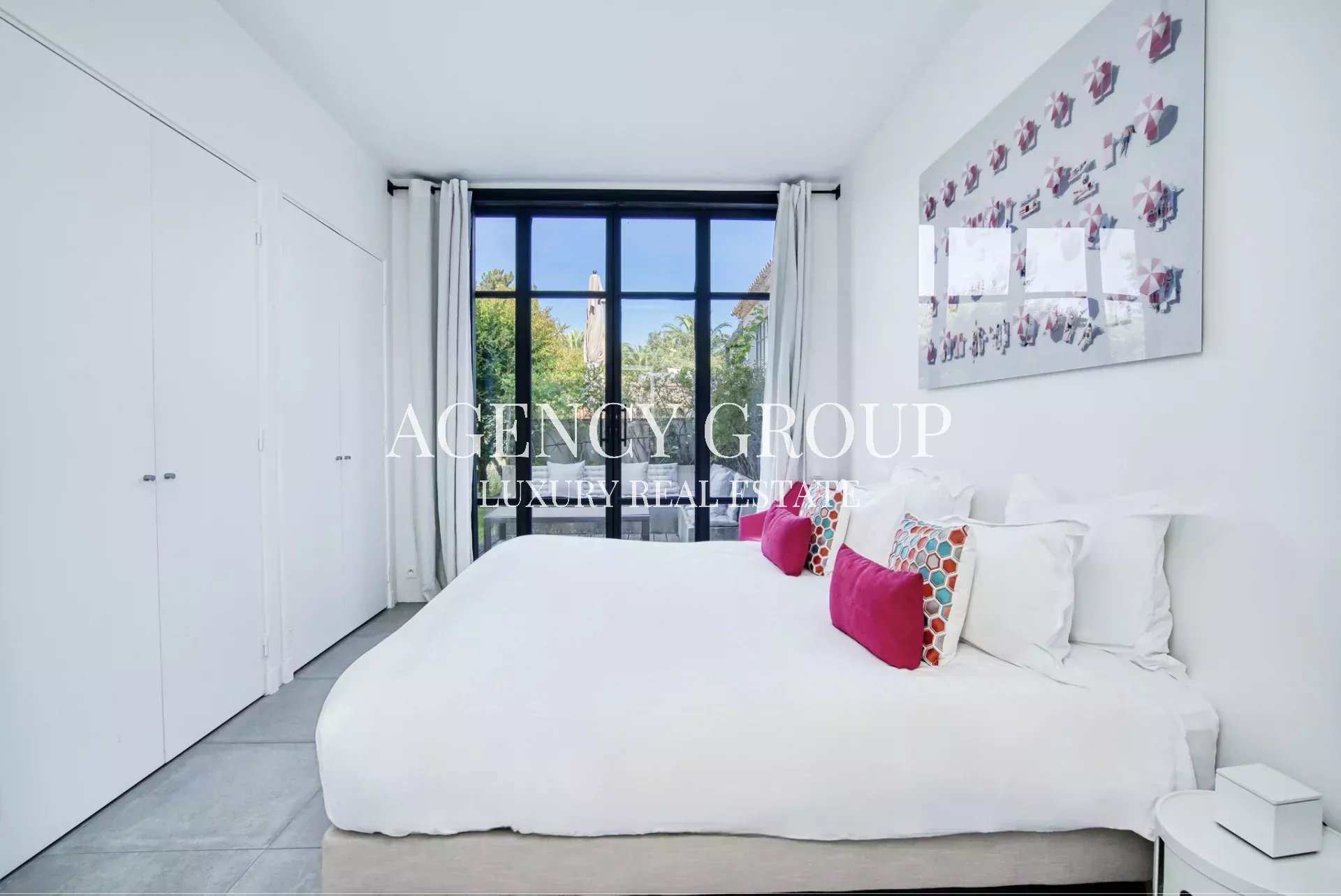 Bright luxury bedroom with a large window, white bed, and colorful throw pillows; minimalist wardrobes along the left wall and abstract art on the right wall.