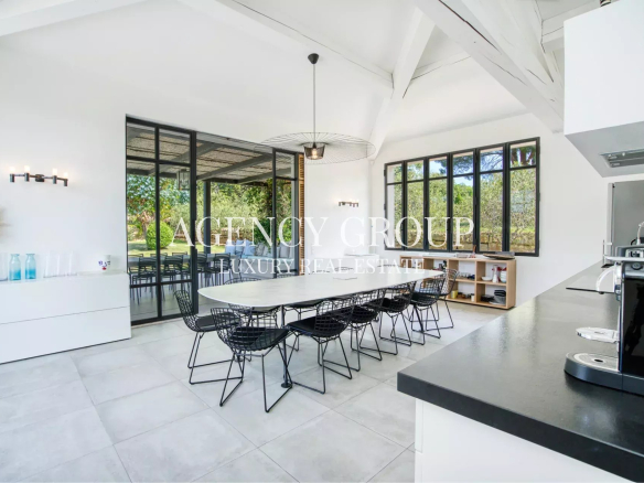 Bright, modern dining area with a long white table, black wire chairs, and large glass doors to an outdoor patio.Kitchen counter and appliances visible on the right; airy, high-ceiling space with white walls.