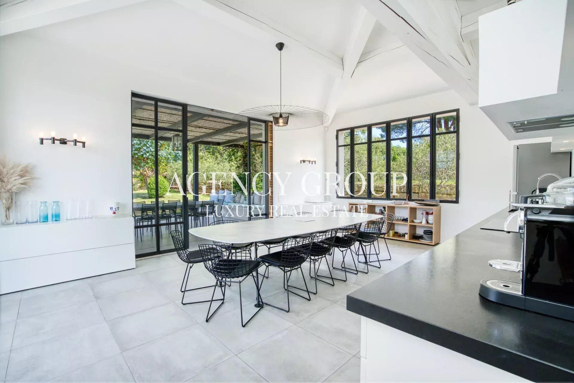 Bright, modern dining area with a long white table, black wire chairs, and large glass doors to an outdoor patio.Kitchen counter and appliances visible on the right; airy, high-ceiling space with white walls.