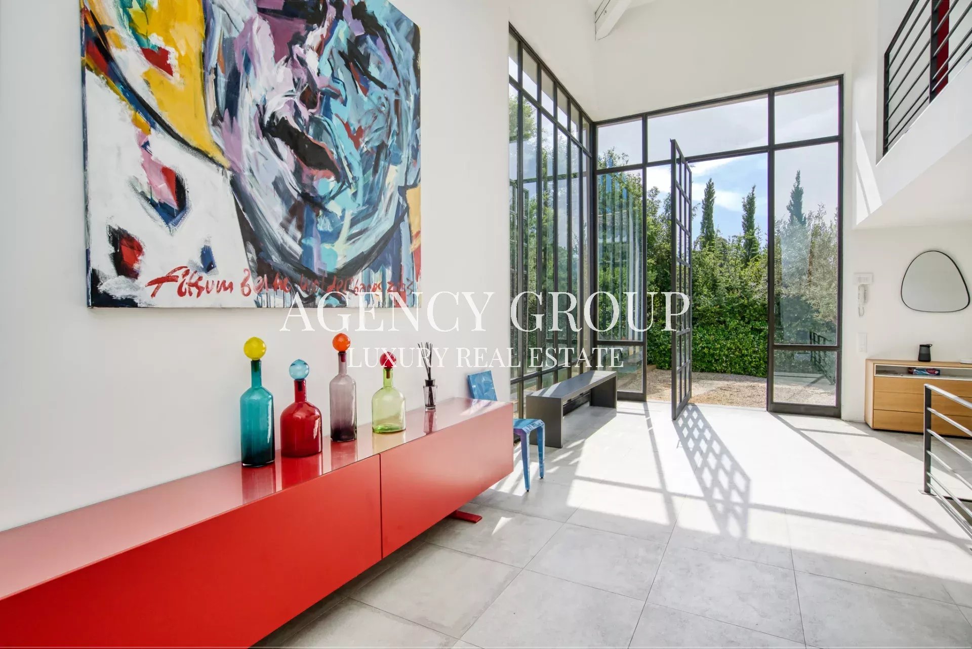 Bright, modern foyer with a large abstract painting on the wall and a red sideboard topped with colorful glass bottles, illuminated by natural light through tall glass doors to a garden.
