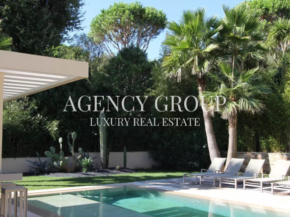 Luxurious backyard pool scene with white lounge chairs, palm trees, and a shaded patio; the branding text 'AGENCY GROUP LUXURY REAL ESTATE' is overlaid across the center.