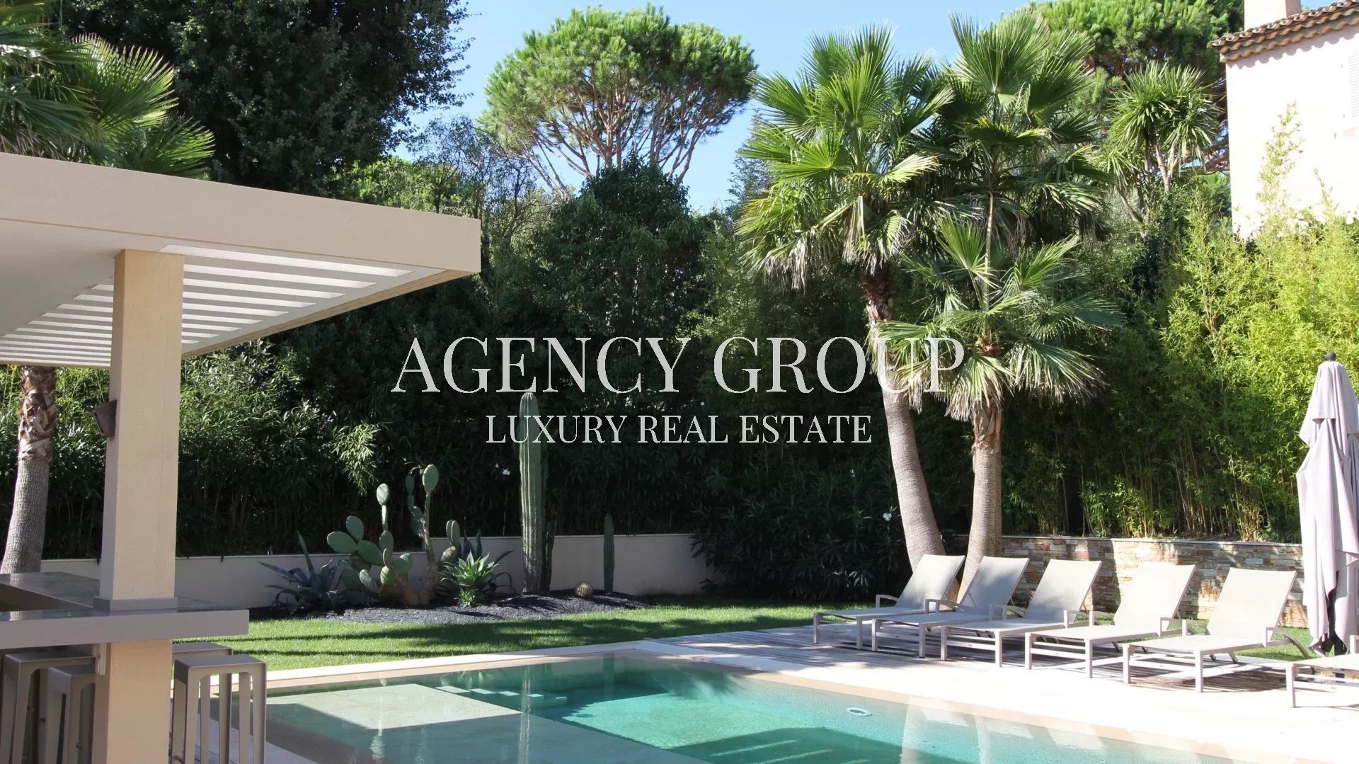 Luxurious backyard pool scene with white lounge chairs, palm trees, and a shaded patio; the branding text 'AGENCY GROUP LUXURY REAL ESTATE' is overlaid across the center.