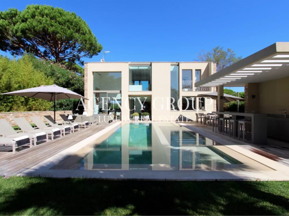 Modern villa with a rectangular pool, loungers, umbrellas, and an outdoor bar under a sunny sky are visible by a glass-walled house.