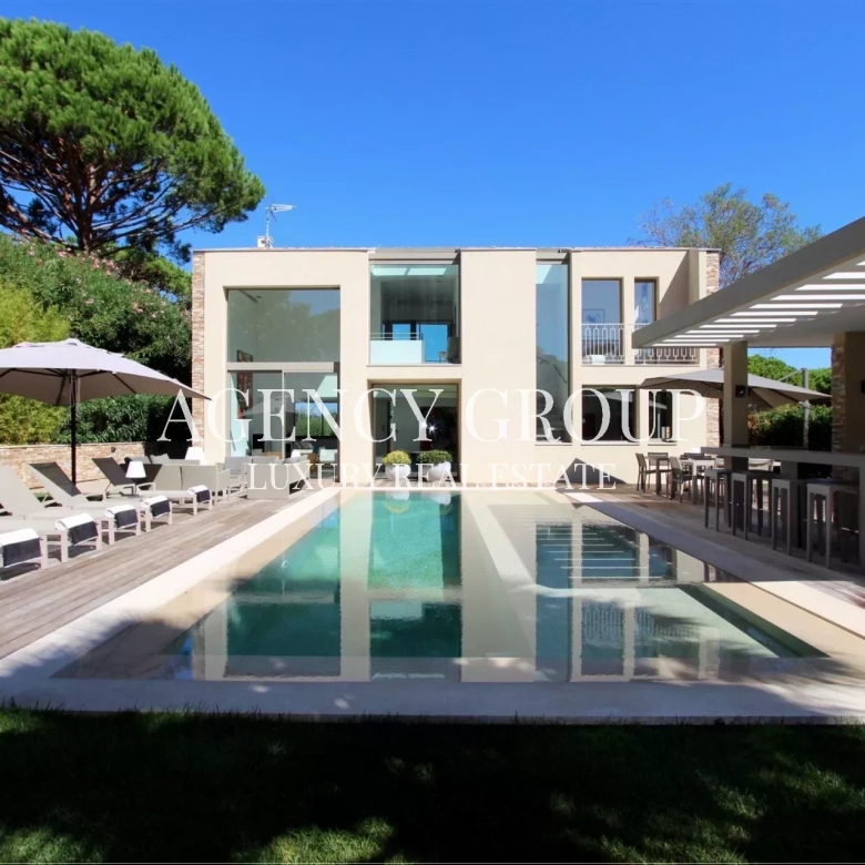Modern villa with a rectangular pool, loungers, umbrellas, and an outdoor bar under a sunny sky are visible by a glass-walled house.