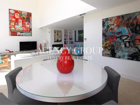 Bright open-plan living and dining area with a round glass table, gray upholstered chairs, and a red vase centerpiece; white walls and bold abstract art framed on the walls.