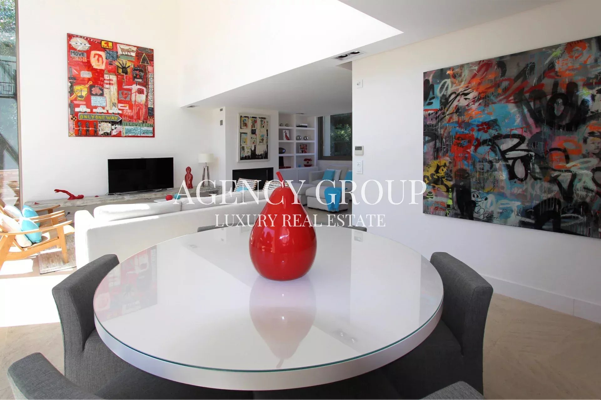 Bright open-plan living and dining area with a round glass table, gray upholstered chairs, and a red vase centerpiece; white walls and bold abstract art framed on the walls.