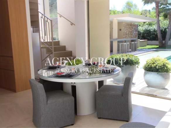 Modern indoor dining area with a round glass table and two gray upholstered chairs, large windows showing a pool and outdoor bar area.