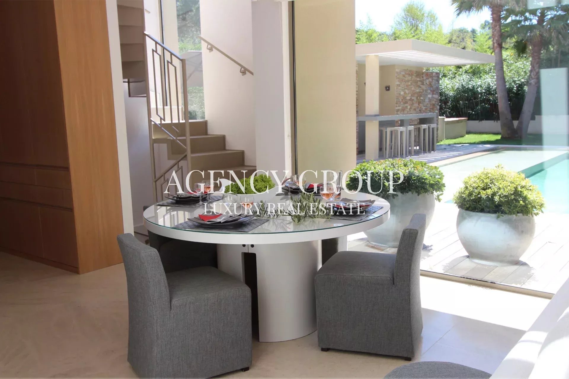 Modern indoor dining area with a round glass table and two gray upholstered chairs, large windows showing a pool and outdoor bar area.