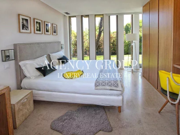 Bright bedroom with a padded headboard bed, white linens, and large floor-to-ceiling windows overlooking a garden.