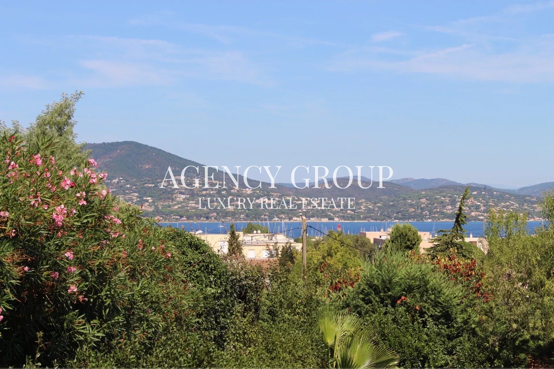 Coastal landscape with hills, blue sea, and green foreground; white overlay text reads 'AGENCY GROUP LUXURY REAL ESTATE'.