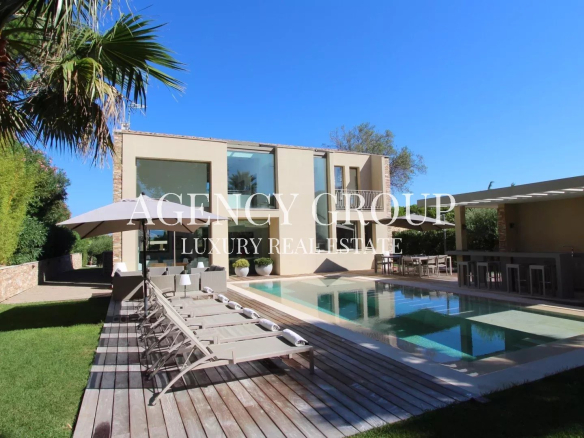 Modern two-story villa with a rectangular pool, wooden deck, sun loungers, and an outdoor dining area under a covered patio.