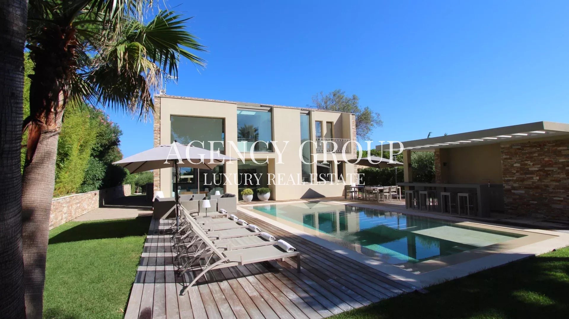 Modern two-story villa with a rectangular pool, wooden deck, sun loungers, and an outdoor dining area under a covered patio.