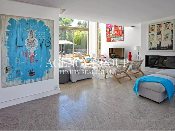 Bright modern living room with white walls, bold blue artwork, glass doors to an outdoor patio, and a seating area with woven chairs and a fireplace.