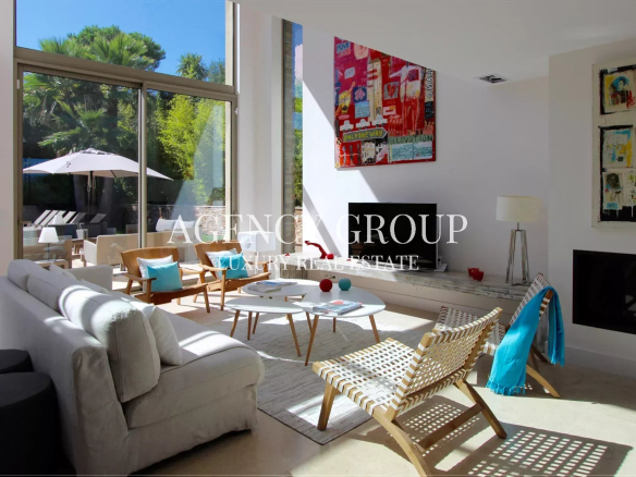 Bright living room with a floor-to-ceiling glass wall opening to a sunny outdoor patio, white sofa and wooden chairs with turquoise cushions
