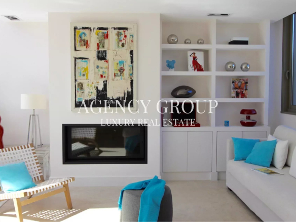 Bright modern living room with white walls, abstract artwork over a gas fireplace, and white shelving filled with decorative items; white sofa with turquoise cushions.