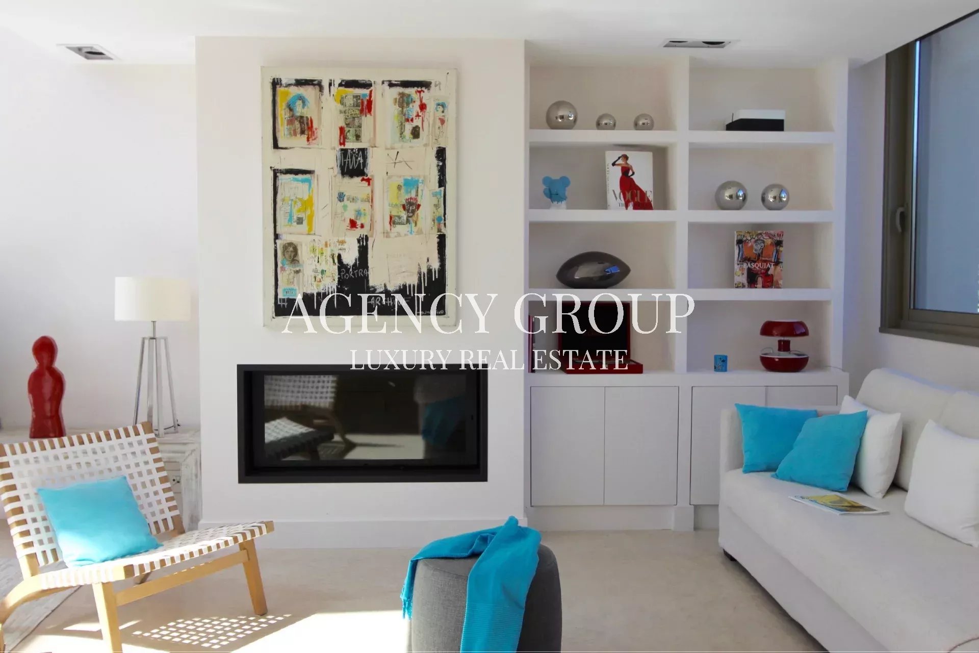 Bright modern living room with white walls, abstract artwork over a gas fireplace, and white shelving filled with decorative items; white sofa with turquoise cushions.