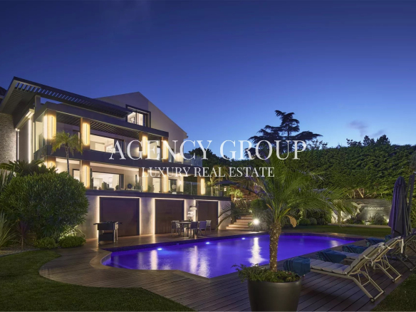 Modern luxury villa at dusk with illuminated exterior, blue-lit pool, and poolside lounge chairs.