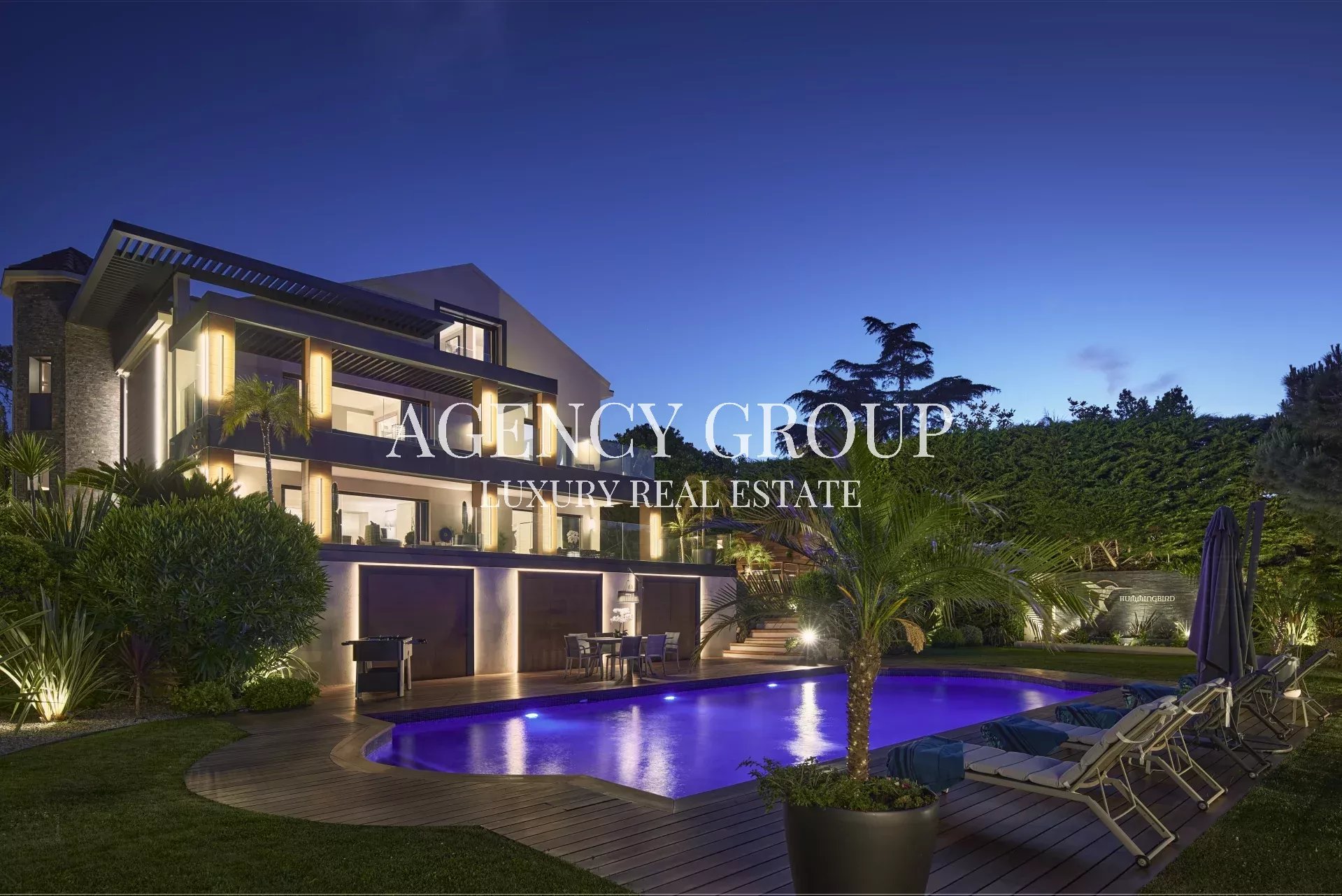 Modern luxury villa at dusk with illuminated exterior, blue-lit pool, and poolside lounge chairs.