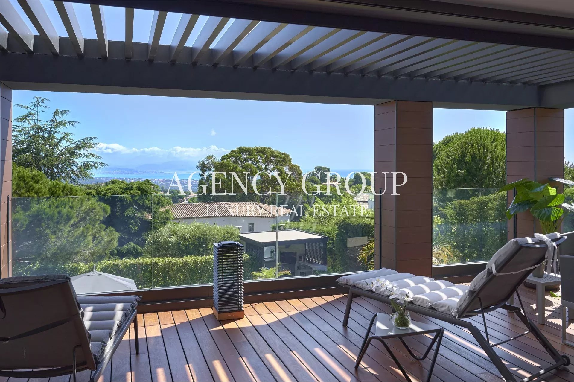 Modern balcony with wooden decking and glass railing overlooking lush trees and distant sea. (watermark: AGENCY GROUP REAL ESTATE)