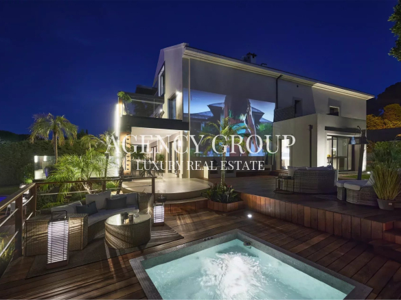 Night view of a modern luxury villa with an illuminated outdoor deck, seating area, and a small pool in the foreground, photographed at dusk.