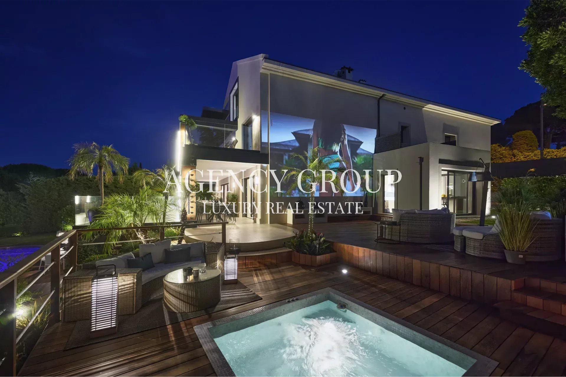 Night view of a modern luxury villa with an illuminated outdoor deck, seating area, and a small pool in the foreground, photographed at dusk.