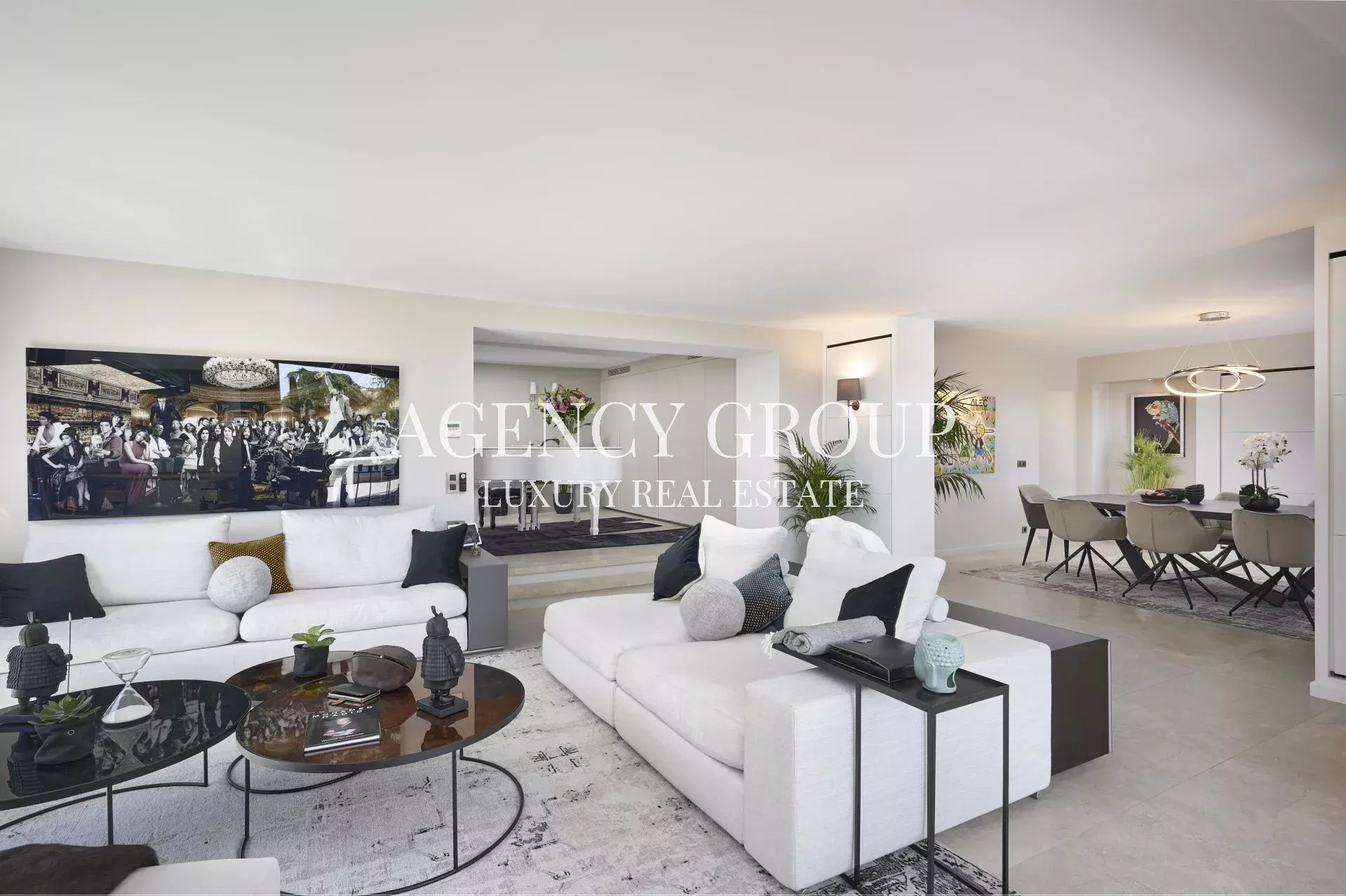 Open-plan living room with white sectional sofas, round glass coffee tables, and neutral decor; dining area visible in the background.