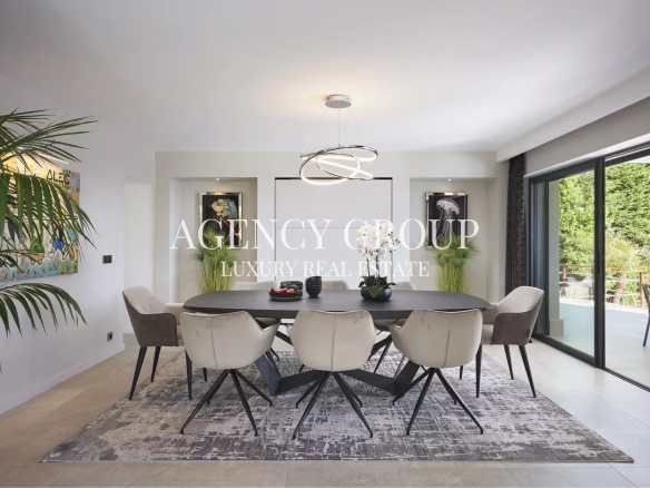 Modern dining room: long black table, eight cream chairs, circular pendant chandelier, and sliding glass doors to a patio with greenery.