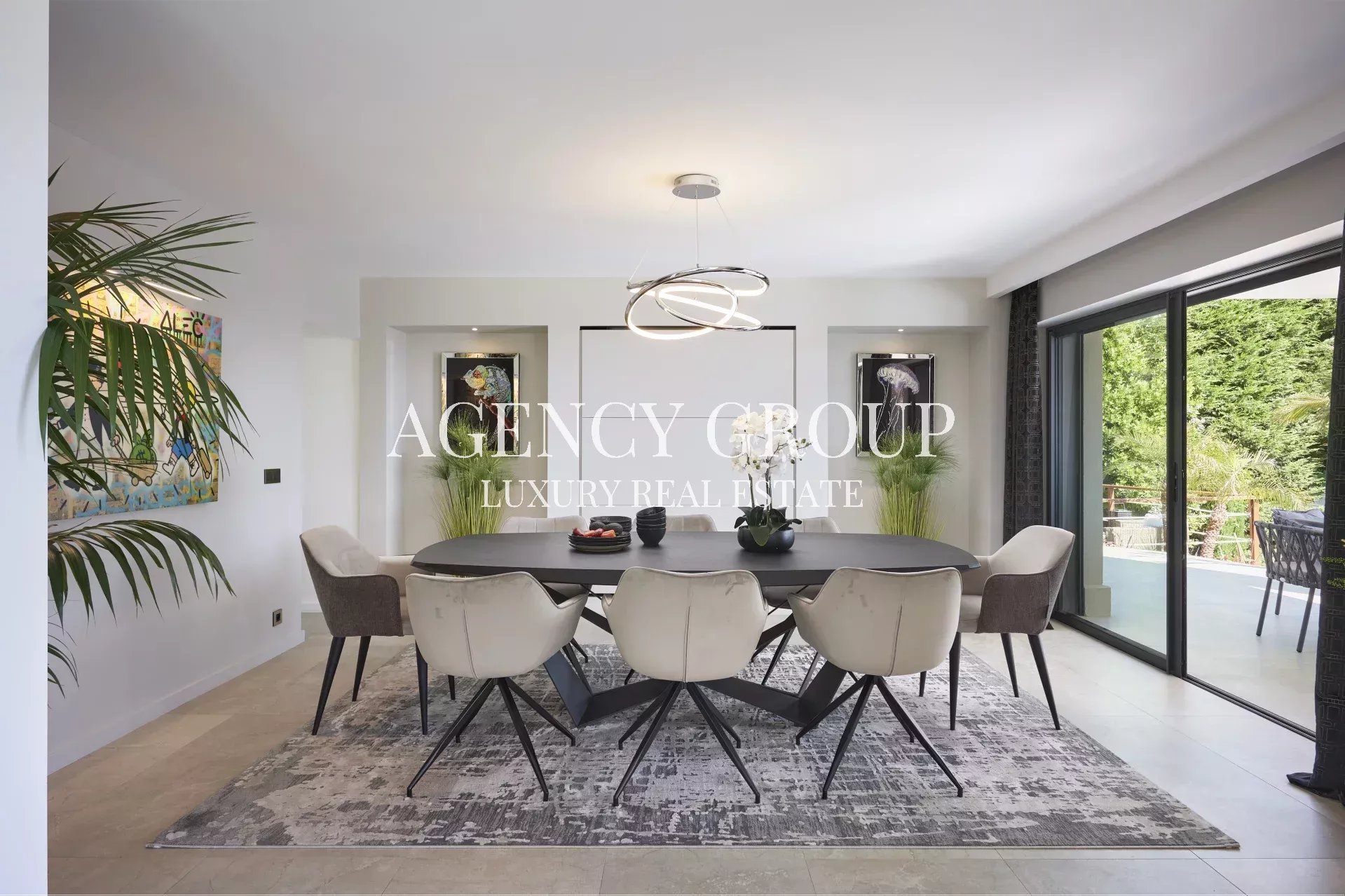 Modern dining room: long black table, eight cream chairs, circular pendant chandelier, and sliding glass doors to a patio with greenery.